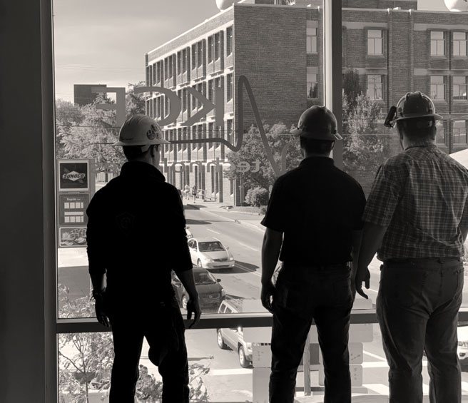 Black and white photo of three men in hardhats looking out a window at the KCF office