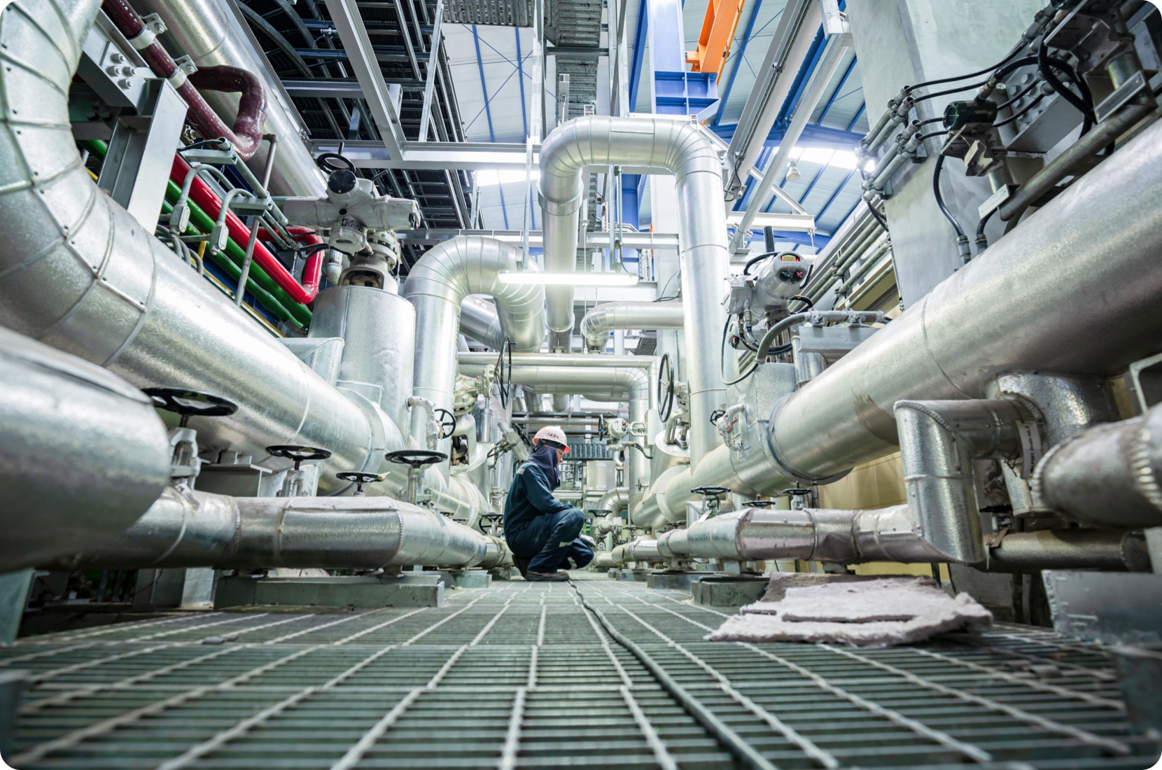Engineer reads machine health data in a plant, surrounded by large pipes and kneeling on a grated floor.