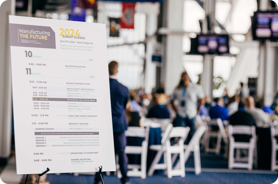 A webinar event sign showing the agenda for the 2024 summit, in the foreground of a blurred event with tables and people attending.